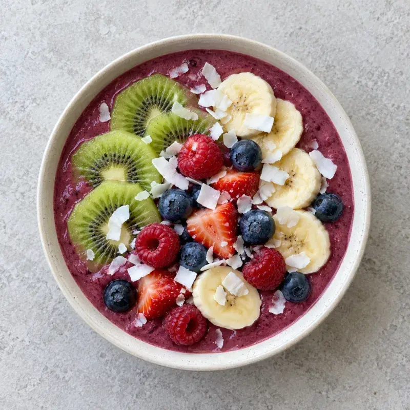 A purple berry smoothie bowl topped with kiwi, banana slices, strawberries, raspberries, blueberries and coconut flakes